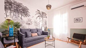 A modern living room with a grey sofa, a small round table, a wicker chair, and a wall mural of palm trees. - Sweett | Rocafort 203 (Barcelona)