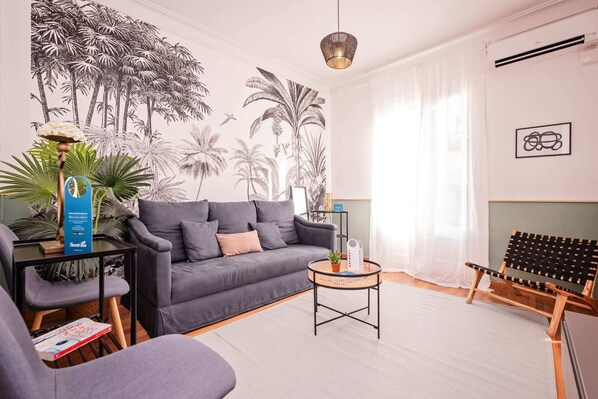 A modern living room with a grey sofa, a small round table, a wicker chair, and a wall mural of palm trees. - Sweett | Rocafort 203 (Barcelona)