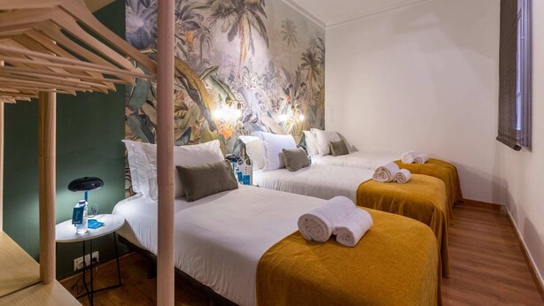 Une chambre d’hôtel avec deux lits, une fresque représentant une végétation tropicale sur le mur, une petite table avec une bouteille d’eau et une fenêtre donnant sur un paysage.