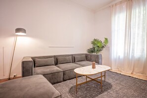 A modern living room with a grey sectional sofa, a small round table, and a floor lamp. - Sweett | Rocafort Principal 3 (Barcelona)
