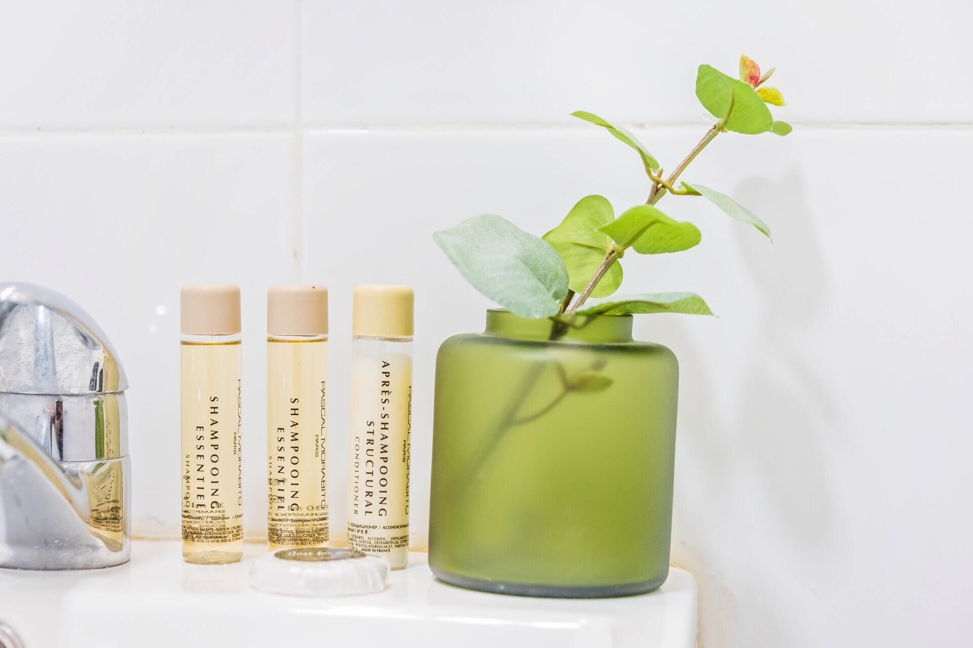 A bathroom with a green vase containing a plant, three bottles of shampoo, and a mirror.