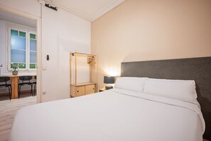A hotel room with a bed, a bedside table with a lamp, a wooden chair, a desk with a vase, and a window with frosted glass. - Sweett | Rocafort Principal 3 (Barcelona)