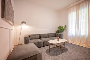 A modern living room with a grey sectional sofa, a small round table, a floor lamp, and a potted plant. - Sweett | Rocafort Principal 3 (Barcelona)