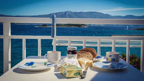 Une table dressée avec des aliments pour le petit-déjeuner, notamment du pain, du fromage et une bouteille, le tout placé devant un paysage pittoresque de mer et de montagnes.