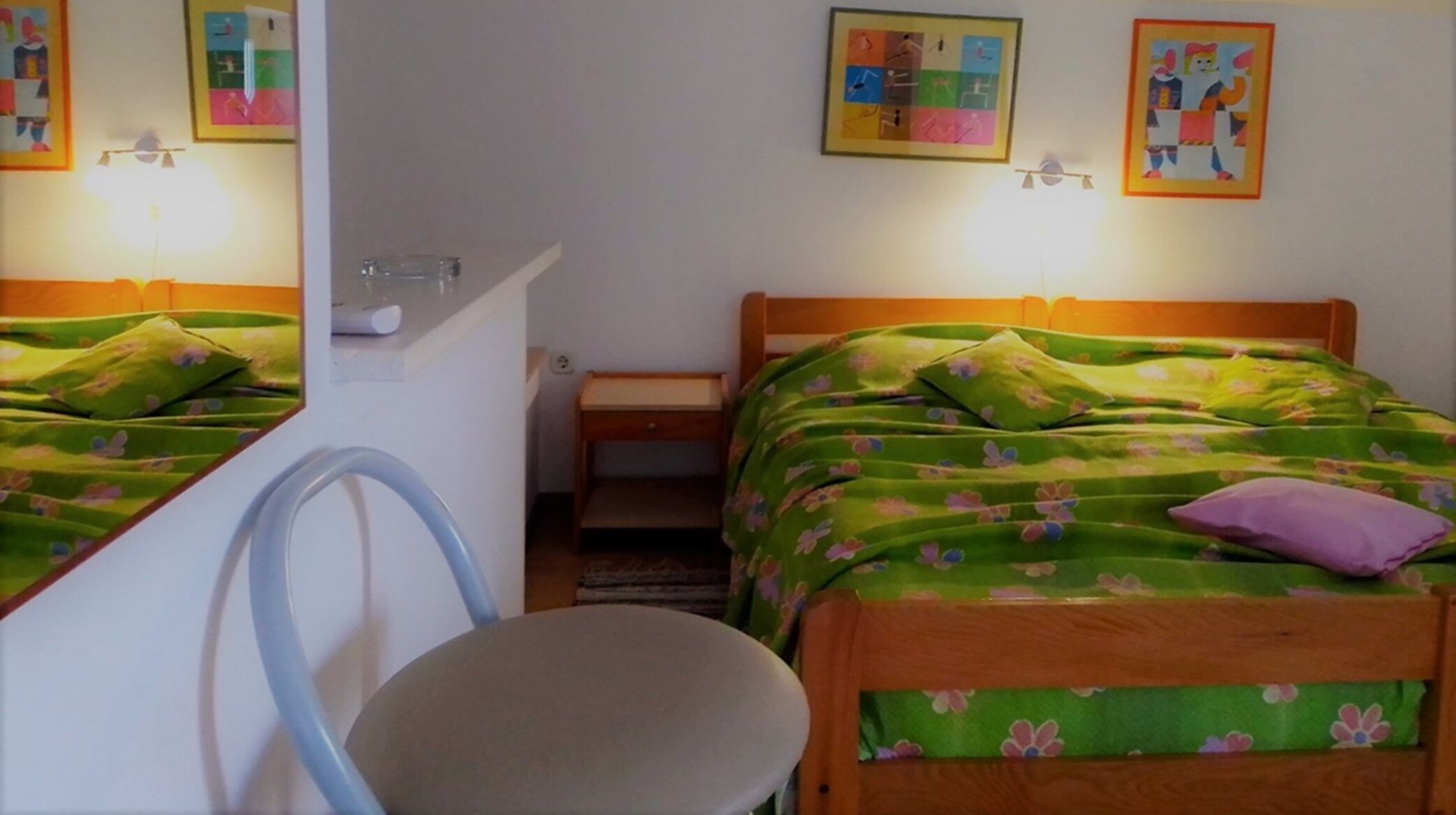 A room with two beds, each with green floral bedding and a wooden headboard. There is a small table and a chair in the room.