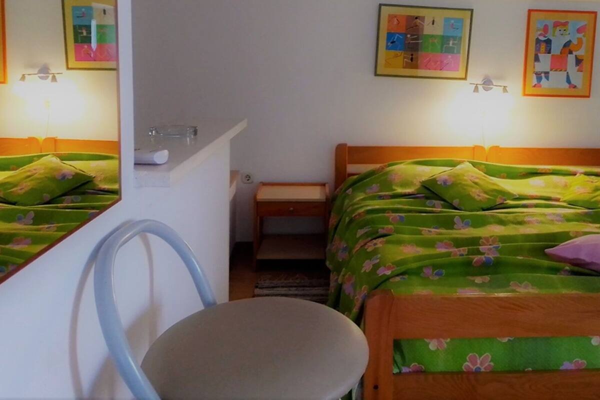 A room with two beds, each with green floral bedding and a wooden headboard. There is a small table and a chair in the room.