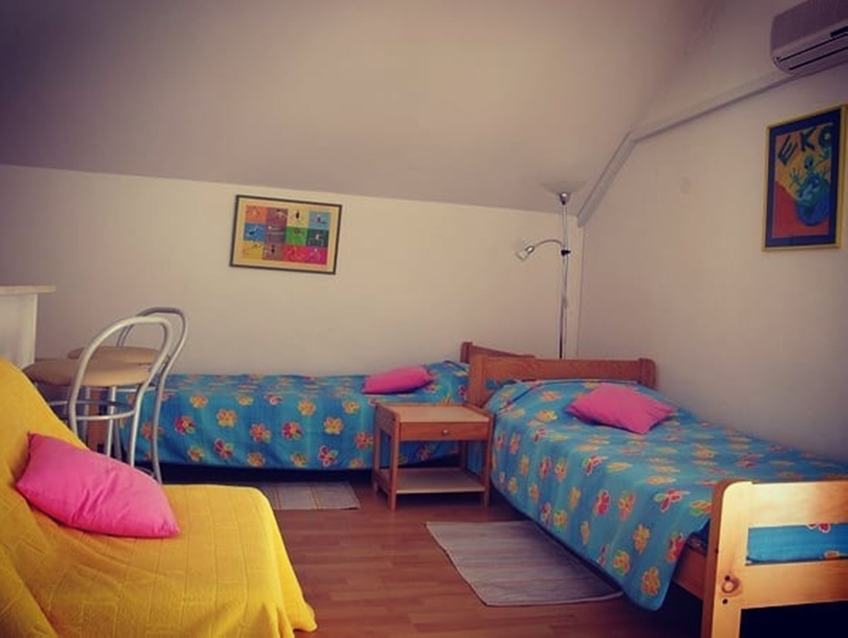 A small, cozy room with two single beds, a chair, a desk, and a wall-mounted air conditioner.