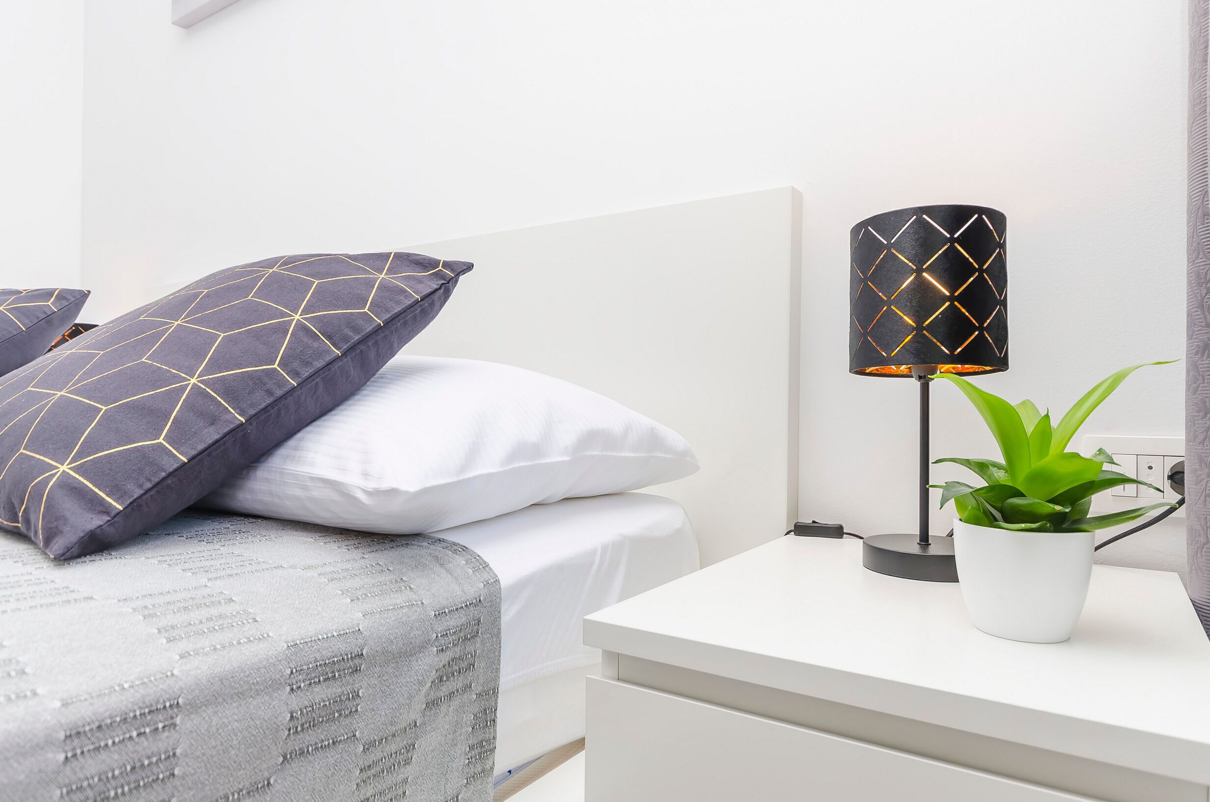 Una cama bien tendida con almohadas estampadas, una lámpara de noche y una planta en maceta sobre una mesita blanca.