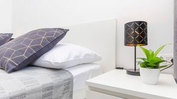 Una cama bien tendida con almohadas estampadas, una lámpara de noche y una planta en maceta sobre una mesita blanca.
