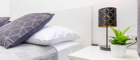 A neatly made bed with patterned pillows, a bedside lamp, and a potted plant on a white nightstand.