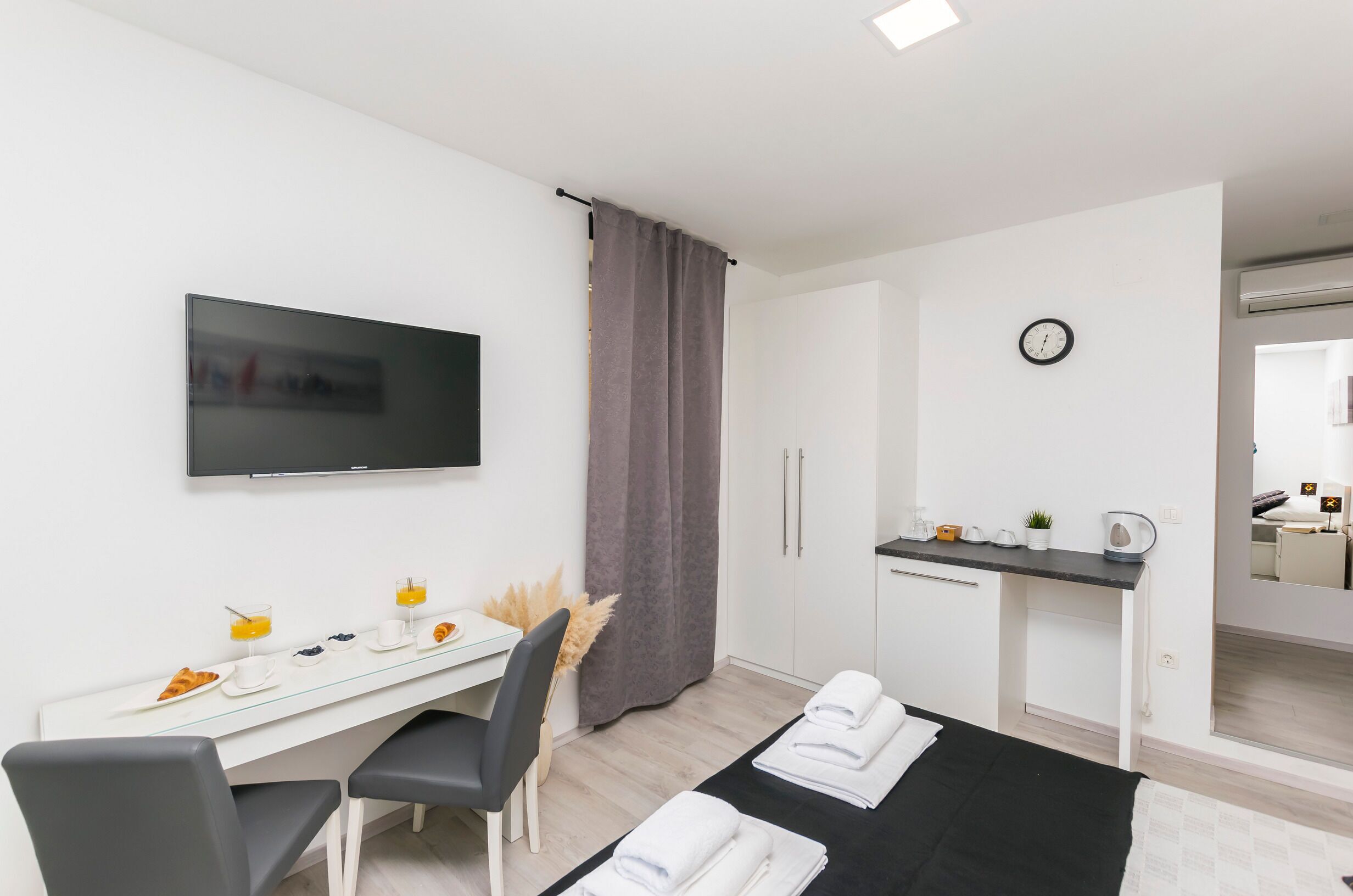 A modern hotel room with a flat-screen TV, a dining area with breakfast, a small kitchenette, and a bathroom visible through an open door.