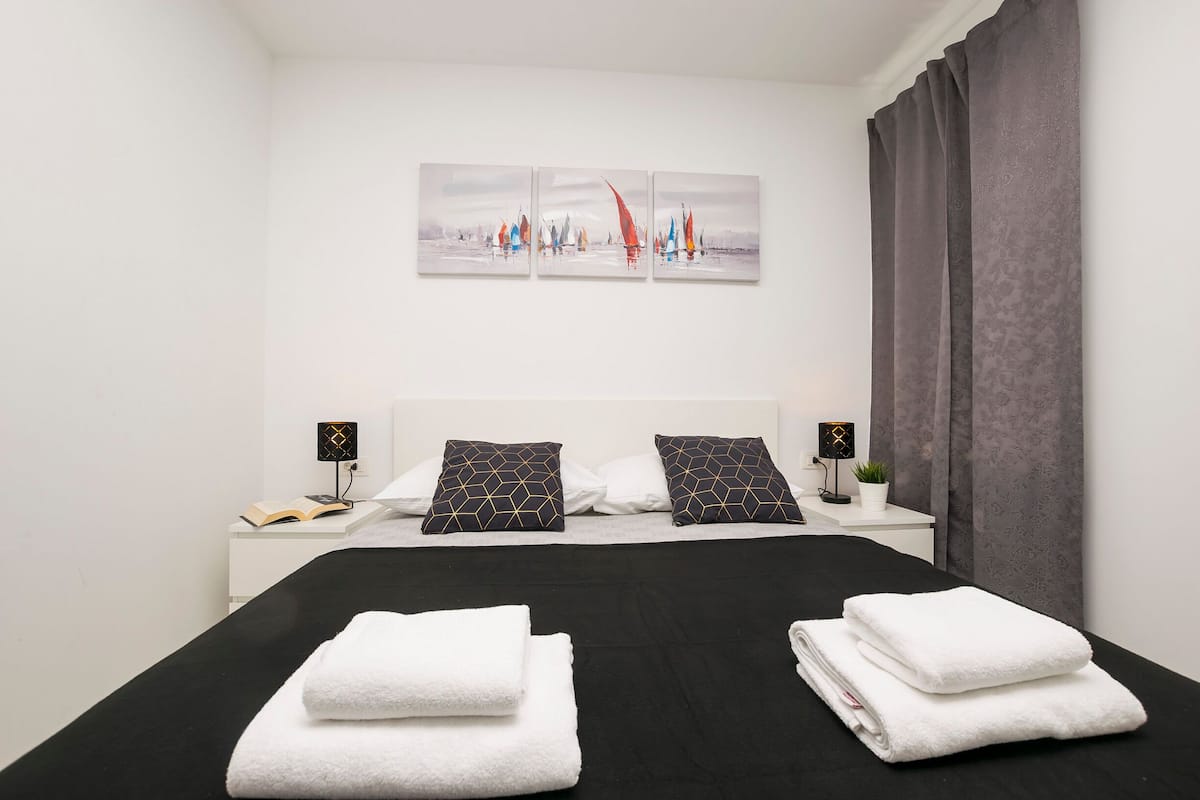 Une chambre d’hôtel comprenant un lit, deux tables de chevet, une commode, une œuvre d’art accrochée au mur, une fenêtre avec des rideaux et une petite plante en pot.