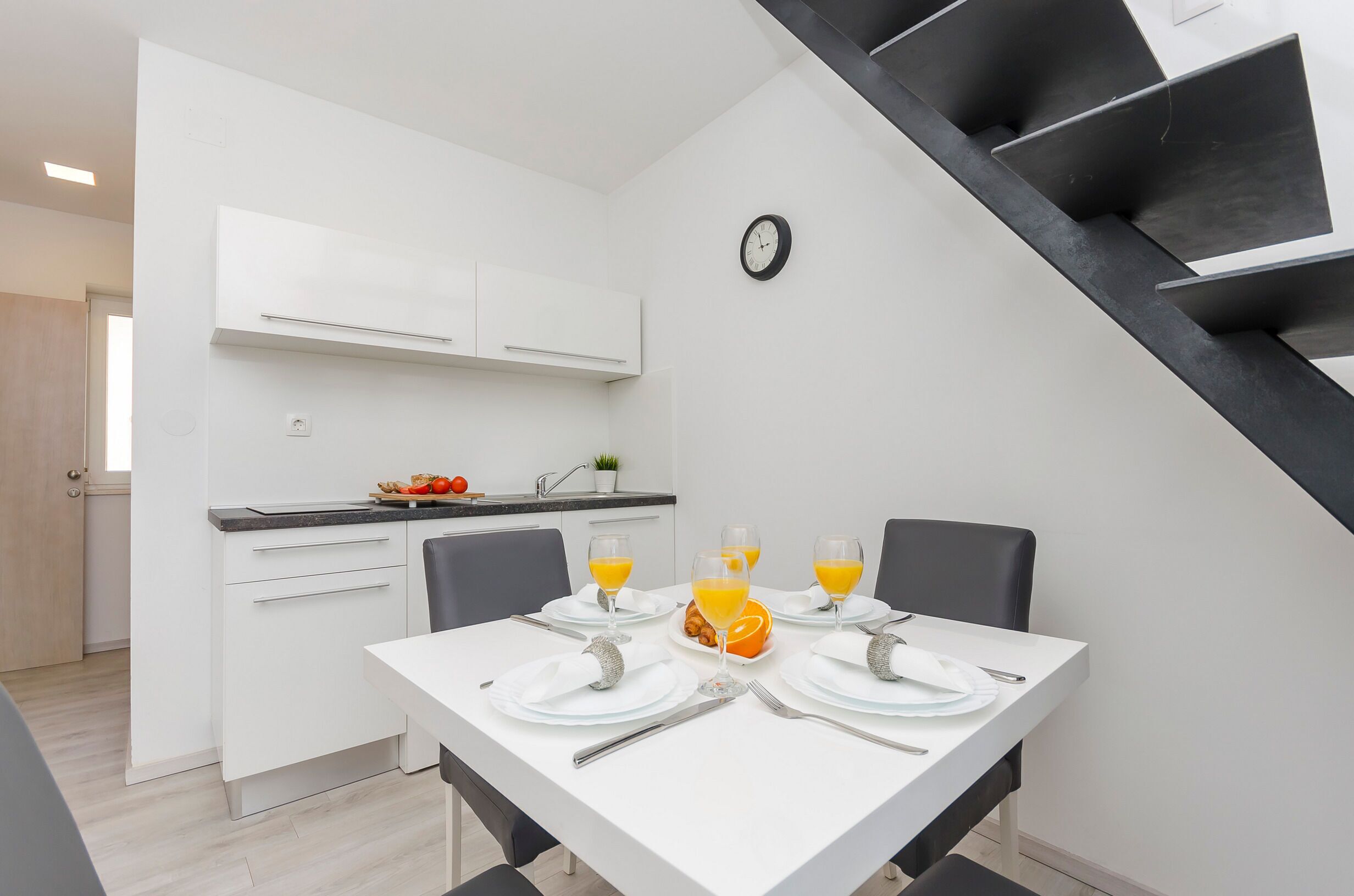 A modern kitchen with white cabinets, a dining table set with plates, glasses, and a fruit bowl.