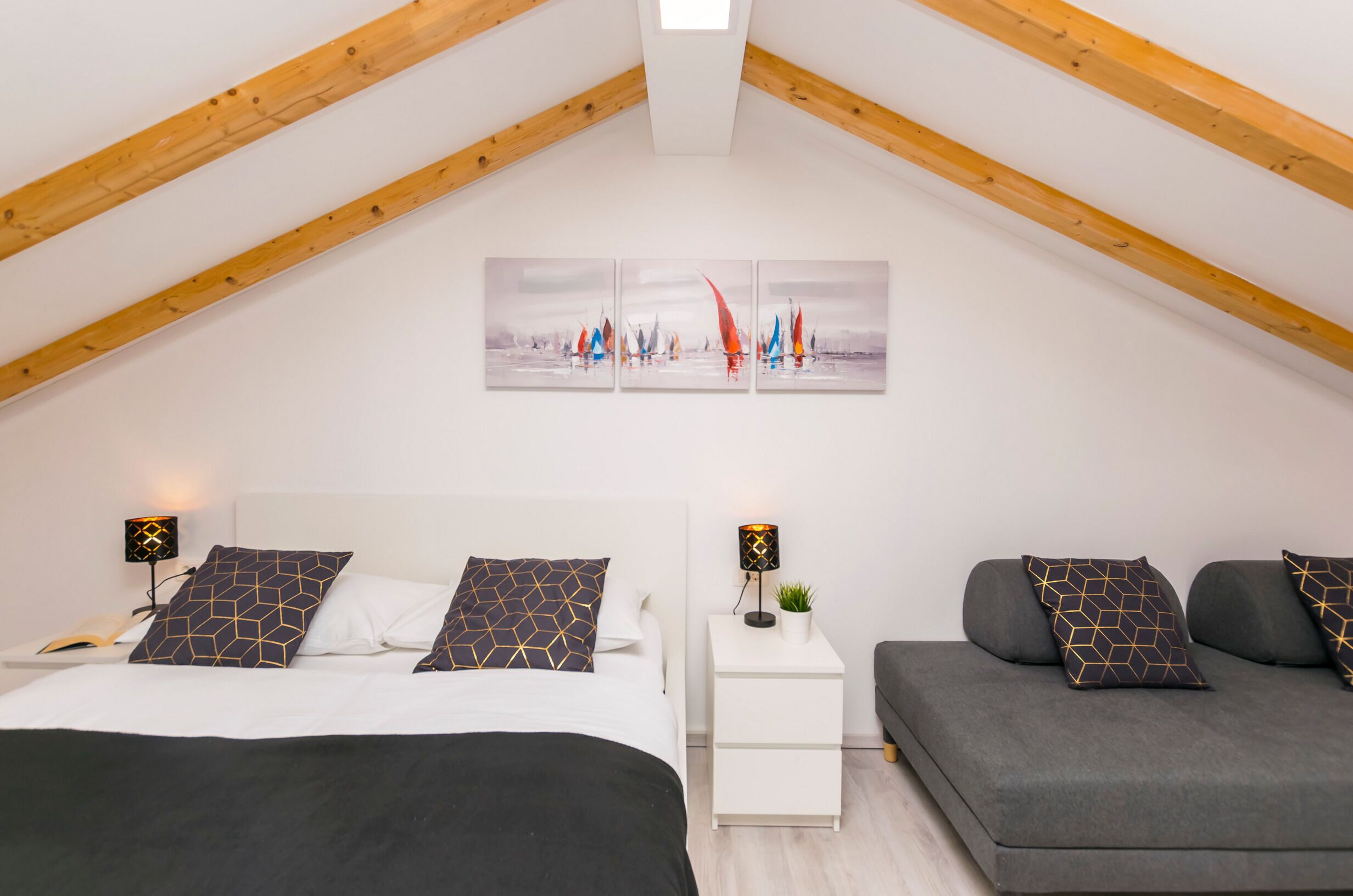 Een moderne slaapkamer met een schuine kap, een bed met gestreepte kussens, een bank met bijpassende kussens en een nachtkastje met een lamp.