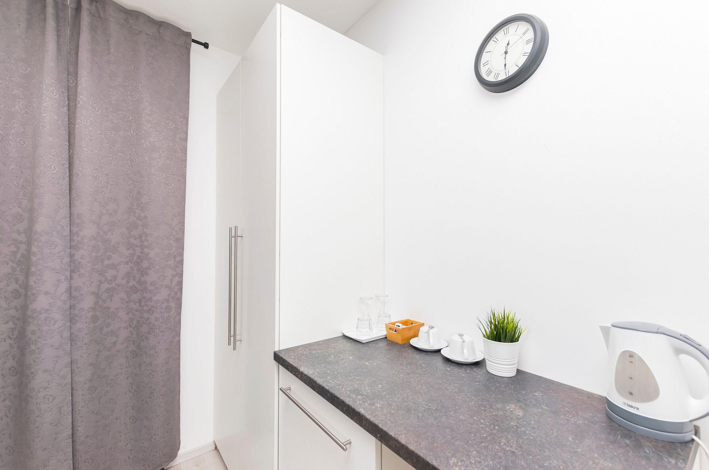 Een moderne keuken met witte kastjes, een grijs aanrecht, een waterkoker, een kamerplant en een klok aan de muur.