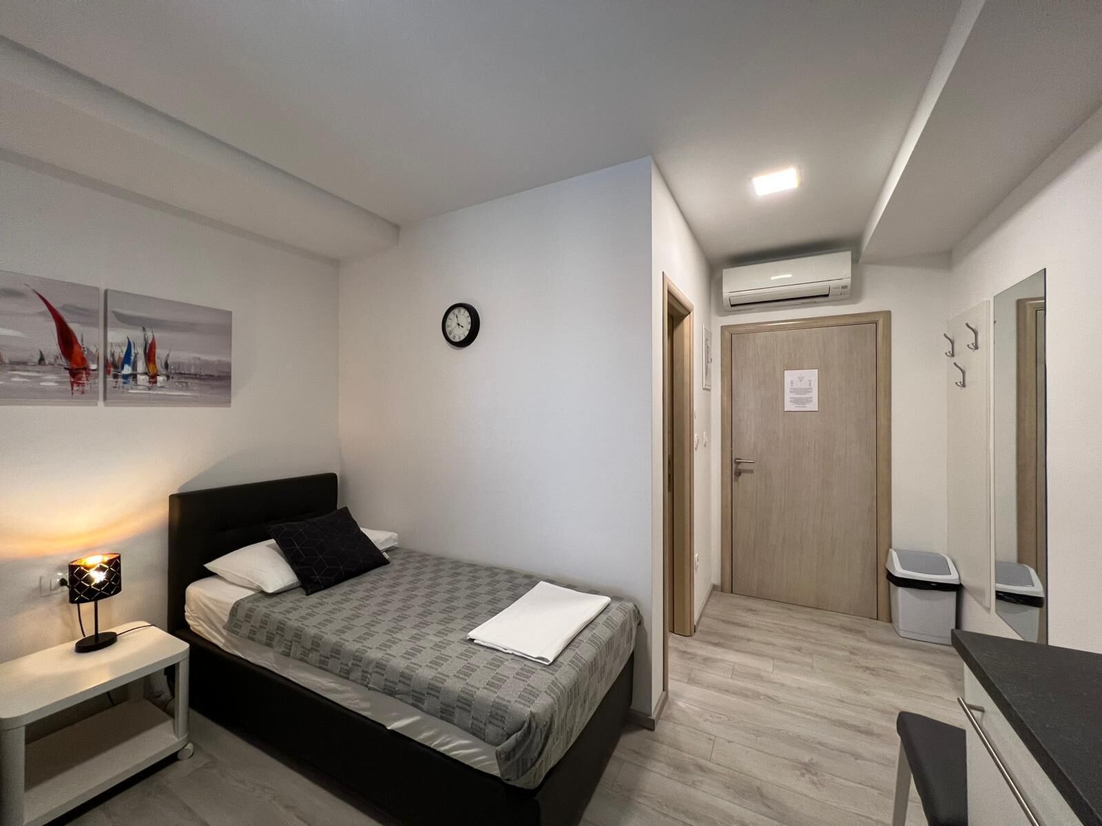 A bedroom with a bed, bedside table, lamp, wall clock, and a door leading to another room.
