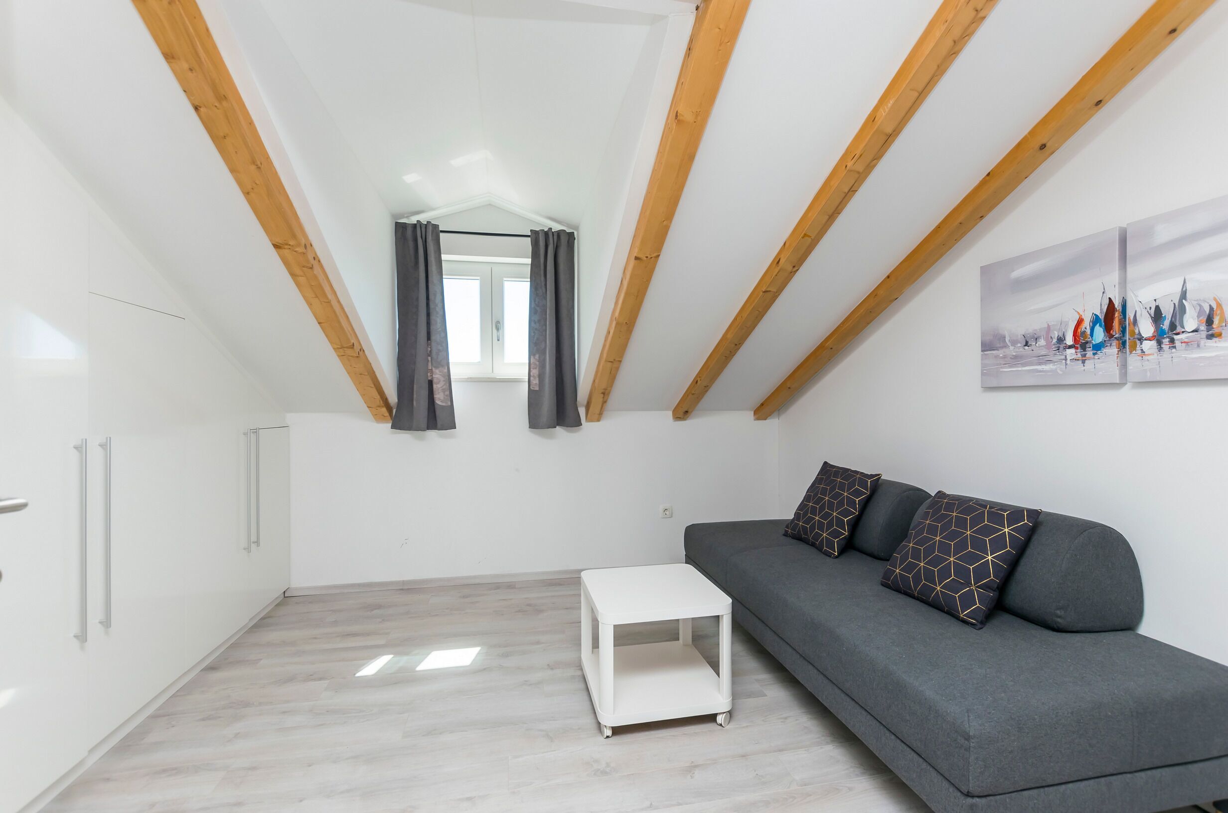 A modern loft bedroom with a sloped ceiling, wooden beams, a gray sofa, a small white side table, and a window with curtains.