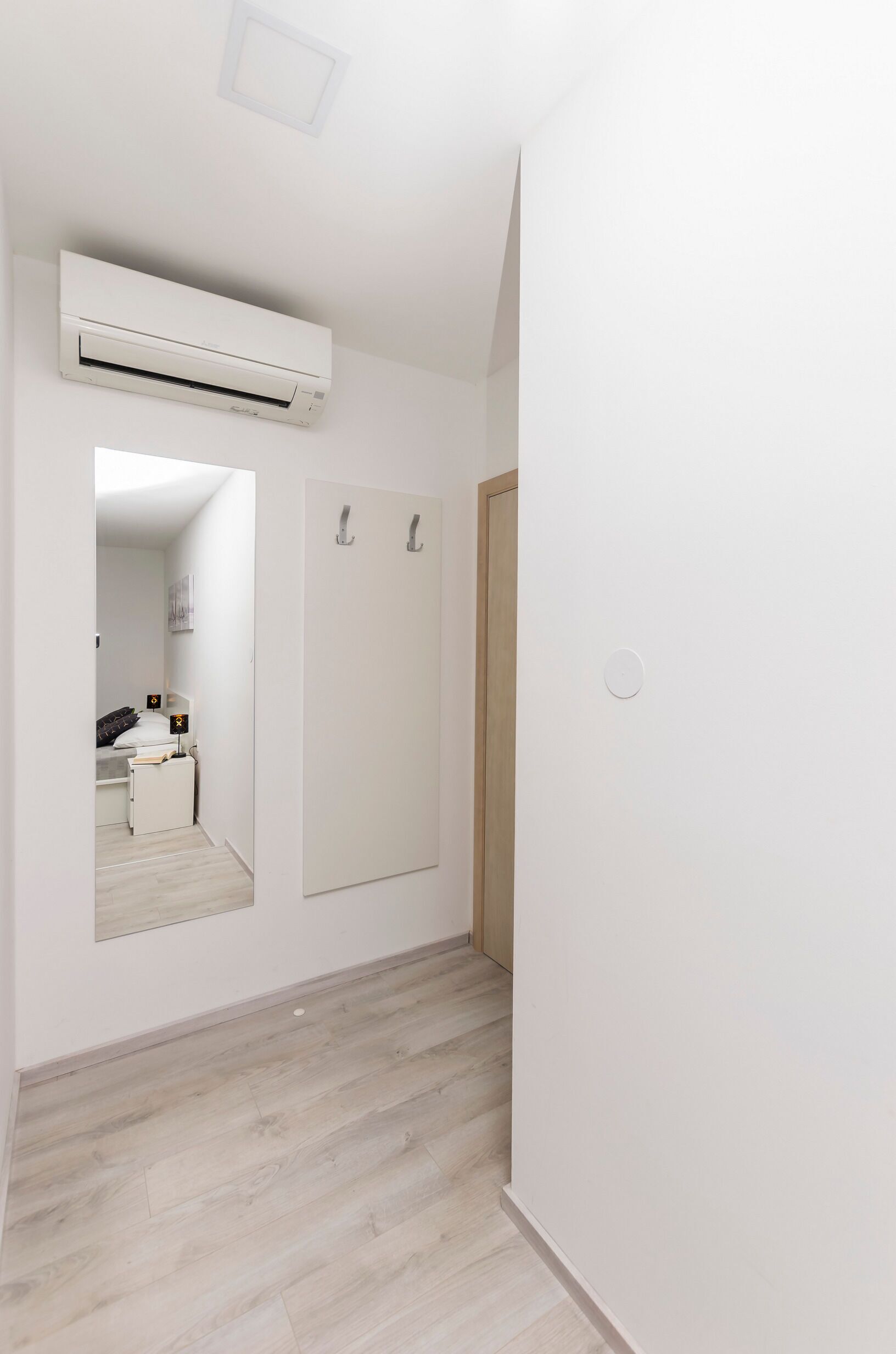 Passadís amb una unitat de aire condicionat blanca, una porta de fusta i un mirall que reflecteix un dormitori amb un llit i una tauleta de nit.