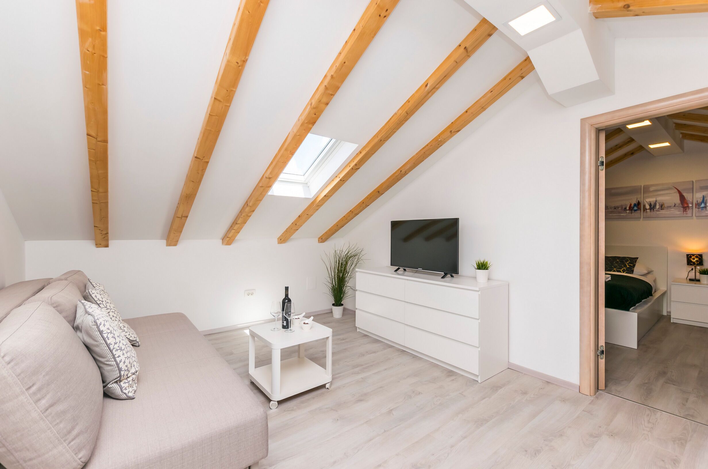 A modern living room with a high ceiling, wooden beams, a flat-screen TV, a sofa, a small white table, and a potted plant.