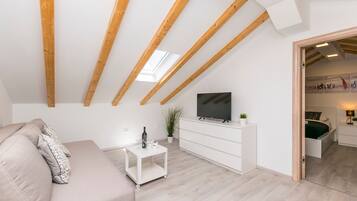 A modern living room with a high ceiling, wooden beams, a flat-screen TV, a sofa, a small white table, and a potted plant.