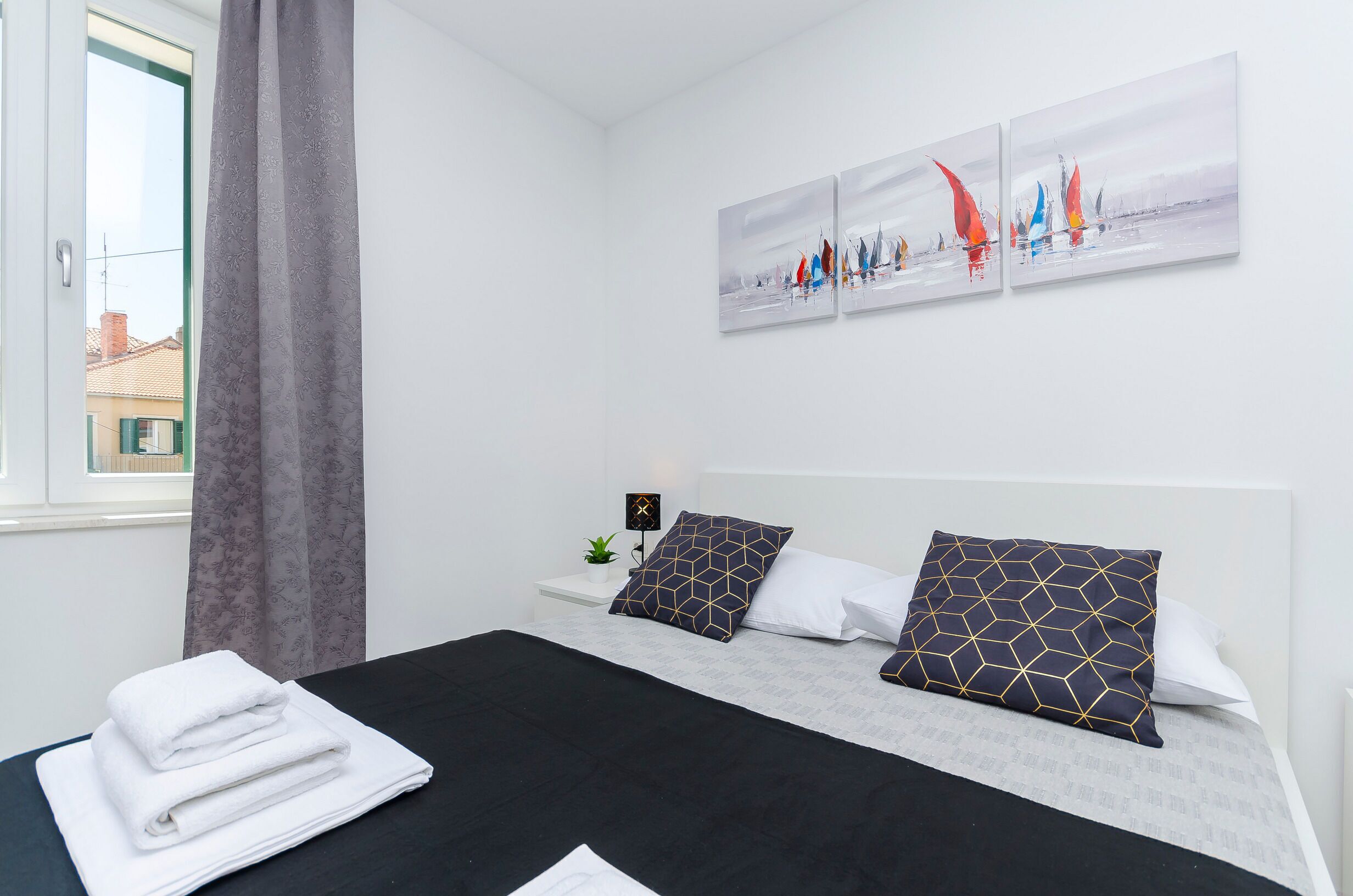 Ein ordentlich gemachtes Bett mit einer schwarz-weißen Tagesdecke, zwei Kissen mit geometrischen Mustern und ein Satz gefalteter weißer Handtücher auf einem Nachttisch.