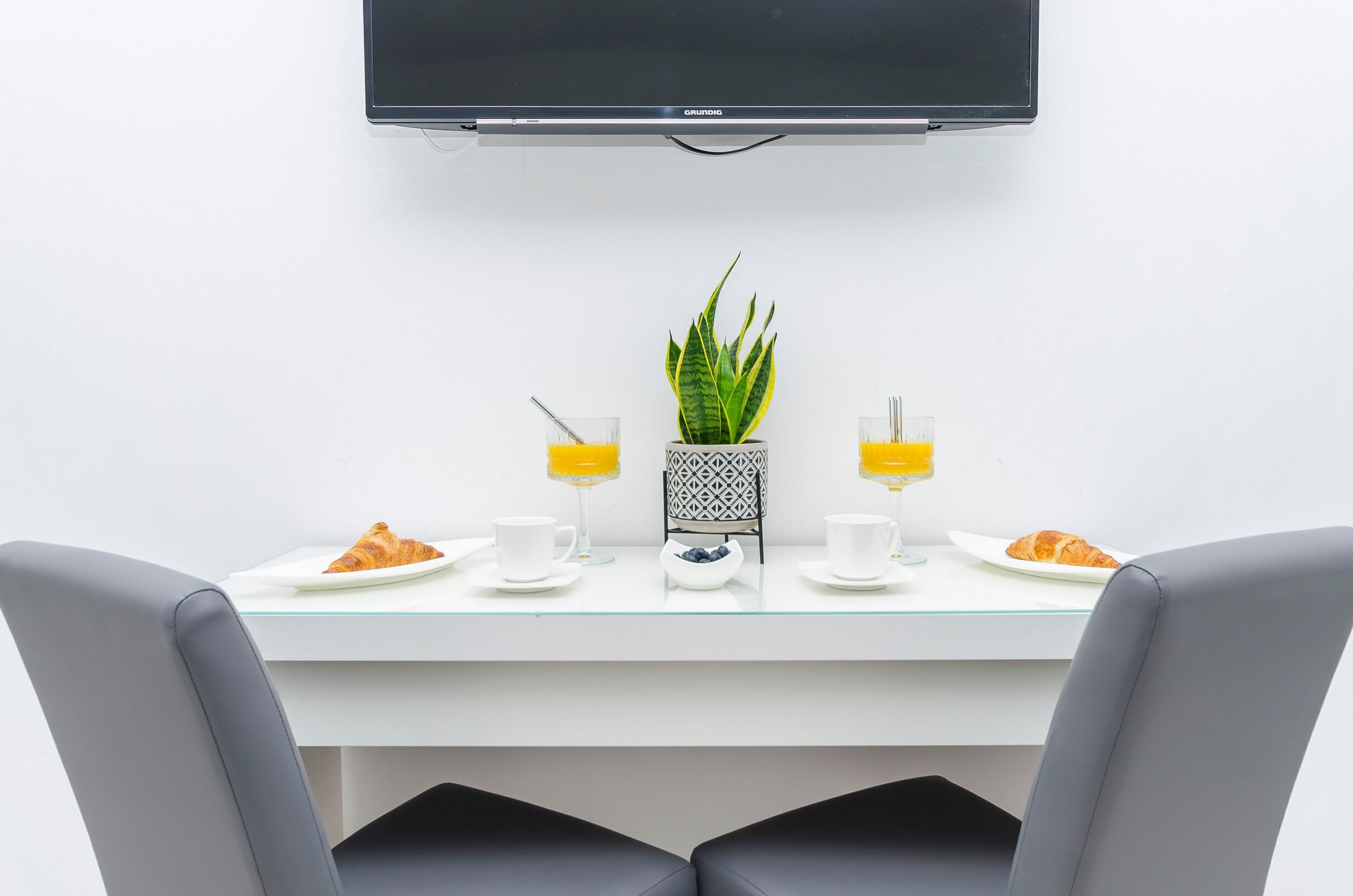 A modern dining area with a flat-screen TV mounted above a white table, two chairs, and a potted plant.