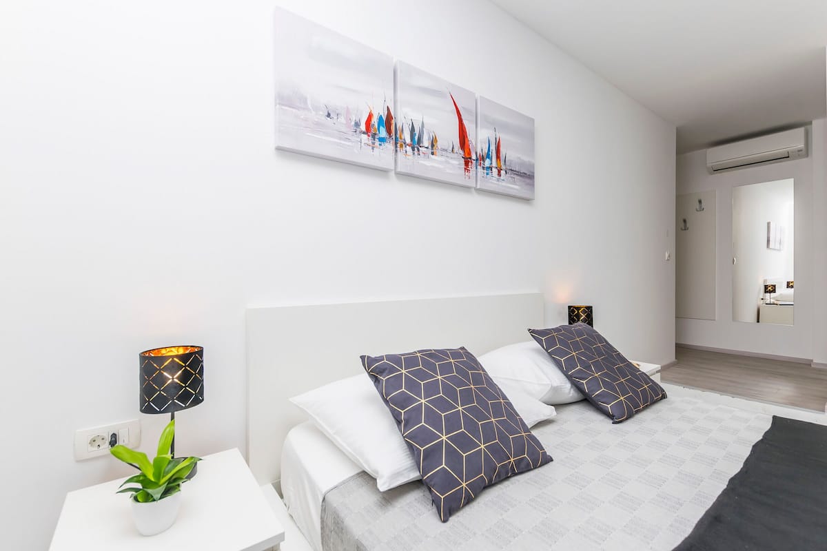 Une chambre moderne avec un grand lit, deux coussins décoratifs, une table de chevet avec une lampe, une plante en pot et une œuvre d’art murale représentant des voiliers.