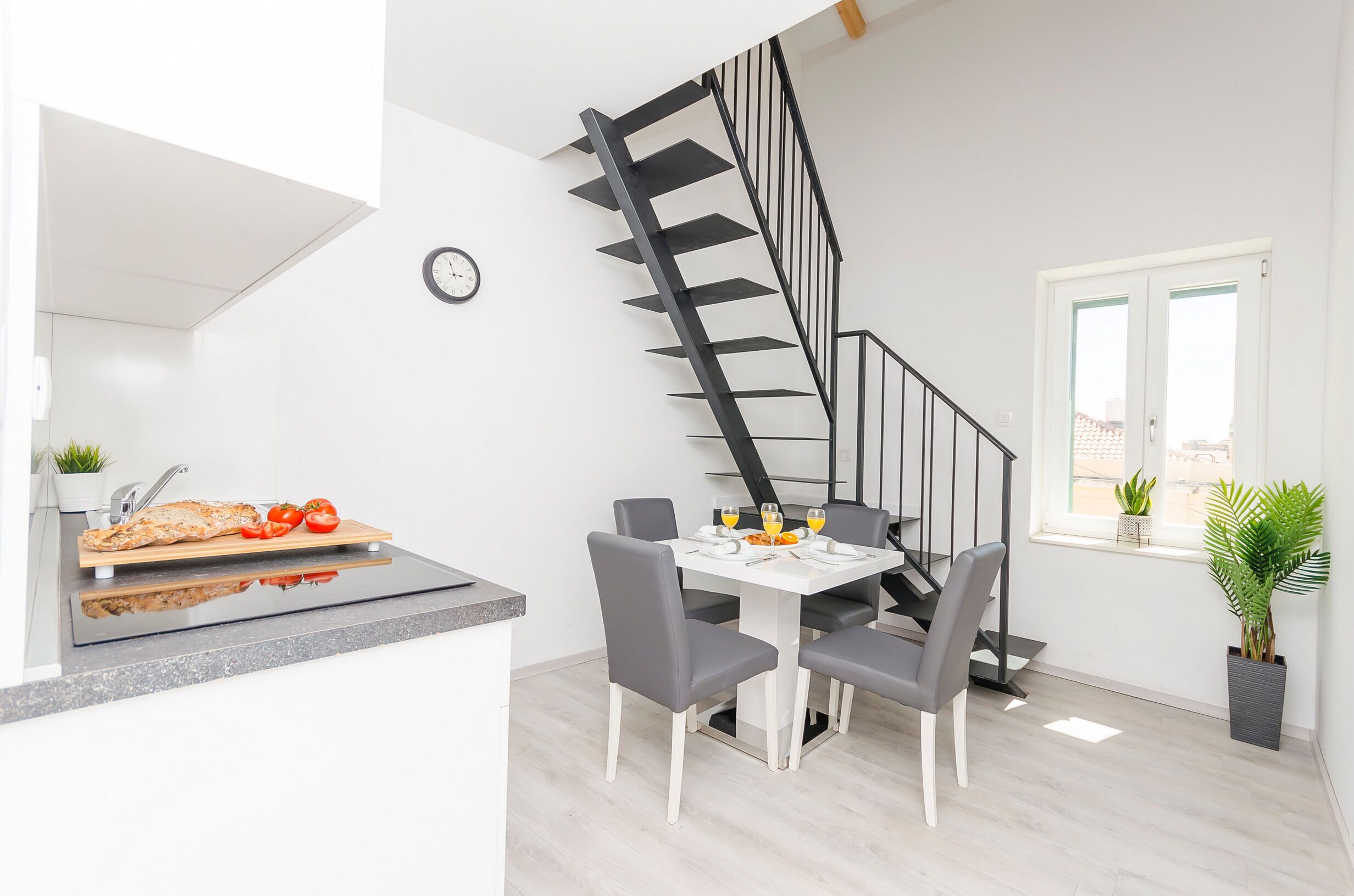 A modern kitchen with a built-in island, a dining area with a white table and grey chairs, and a black spiral staircase.