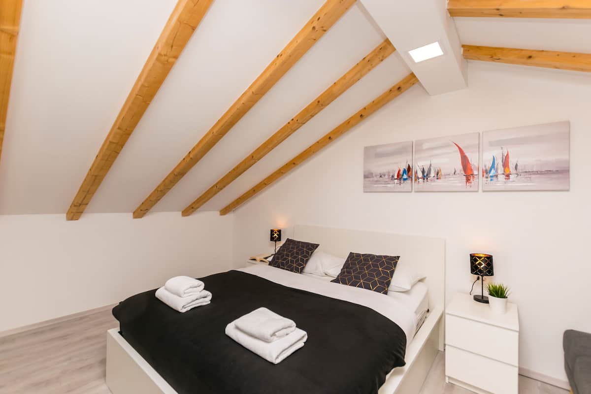 Une chambre moderne avec un plafond en pente, un lit sur lequel sont soigneusement pliées des serviettes, une table de chevet avec une lampe, et un mur orné de trois tableaux.