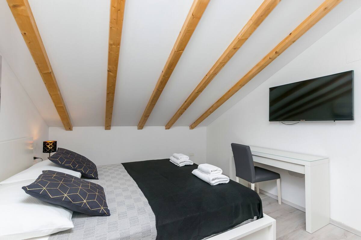 Une chambre moderne avec un lit, deux oreillers, une couverture noire, un bureau, une chaise et un téléviseur fixé au mur.