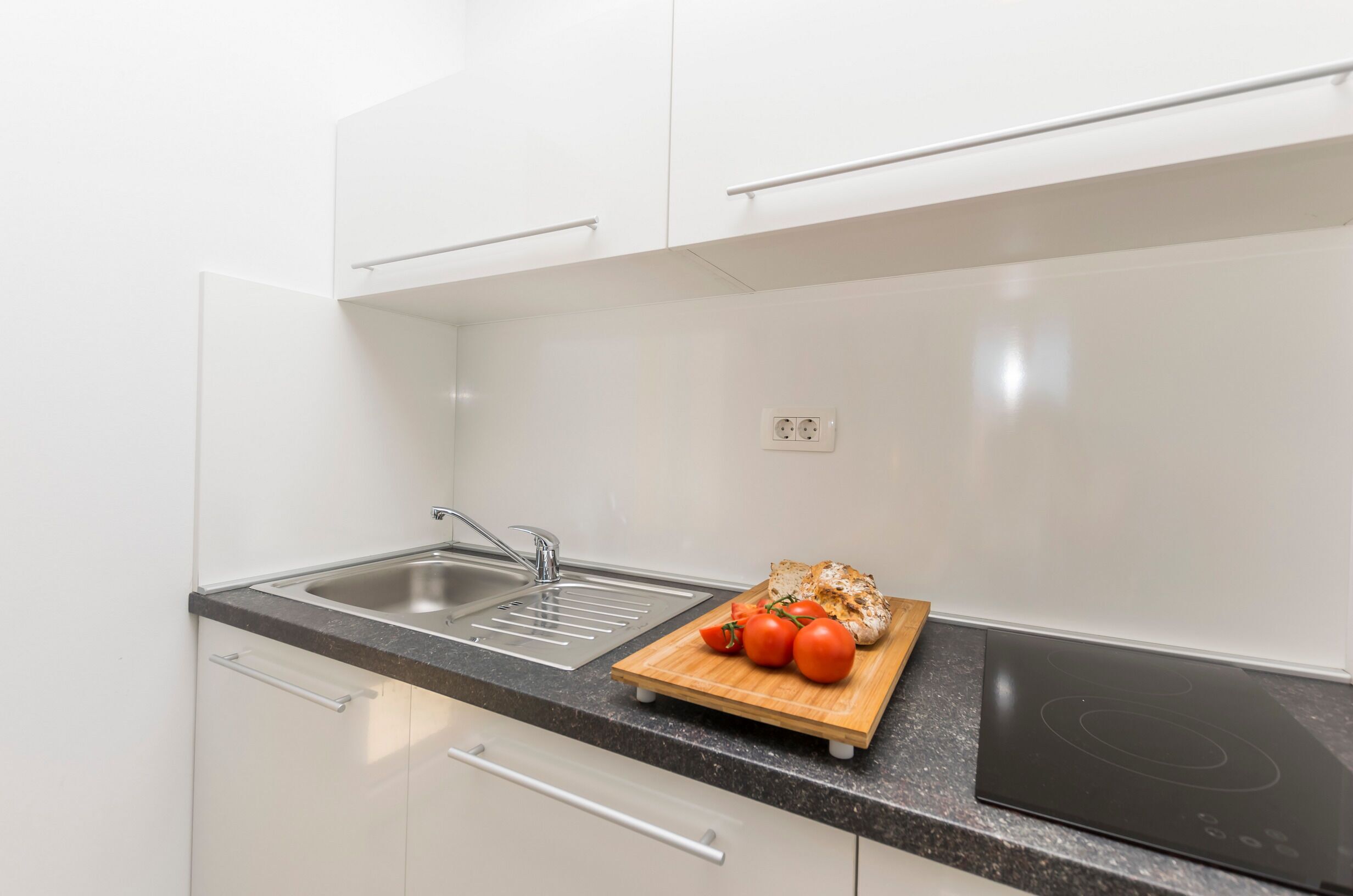 A modern kitchen with white cabinets, a black countertop, a stainless steel sink, and a wooden cutting board with tomatoes and bread.
