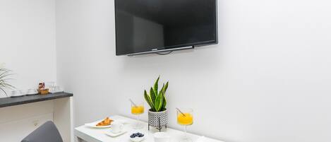 Una habitación moderna con una televisión de pantalla plana en la pared, un escritorio blanco con productos de desayuno, una silla y una planta en maceta.