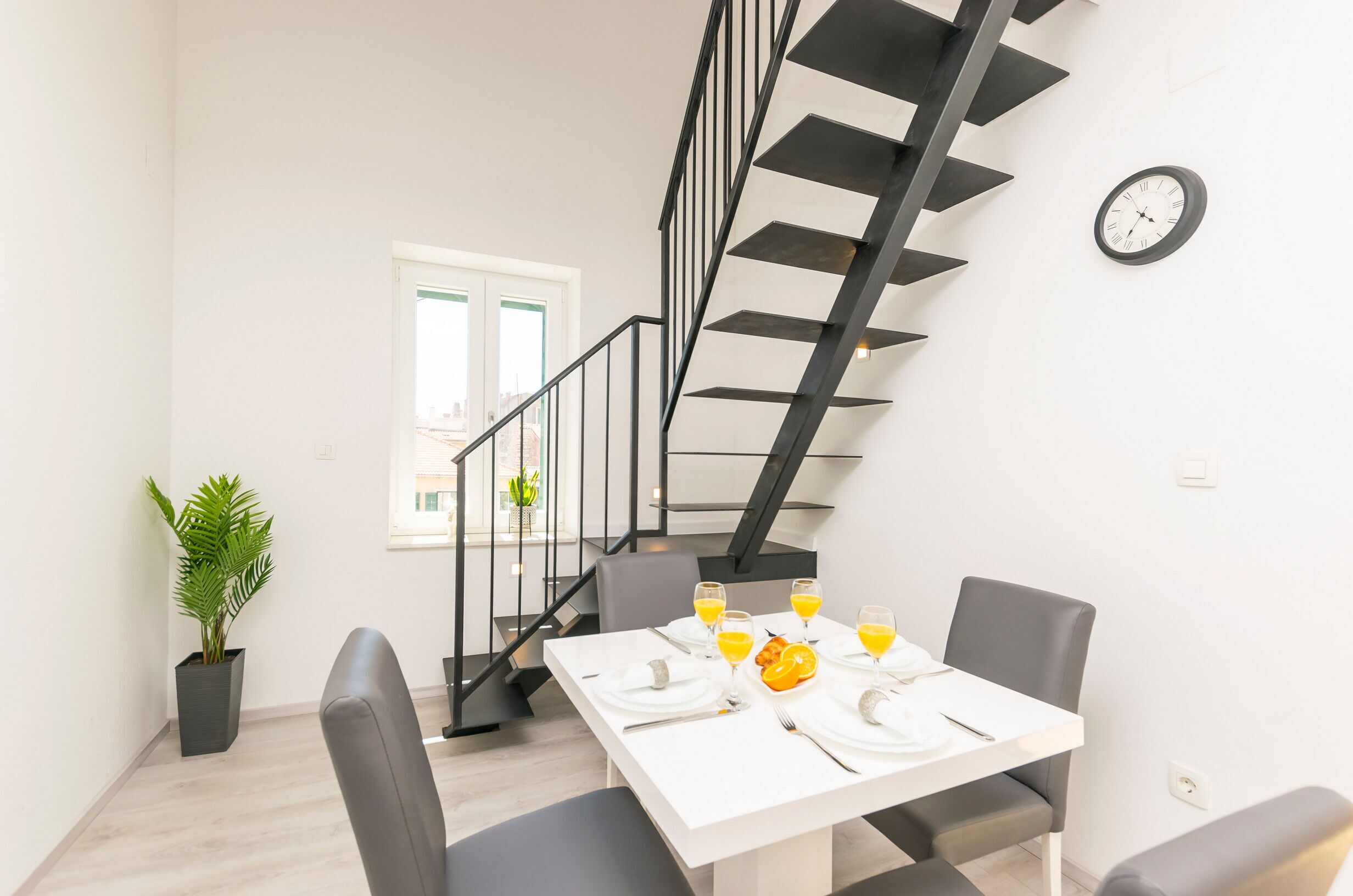 A dining area with a white table set for four, a potted plant, and a staircase with black railings.