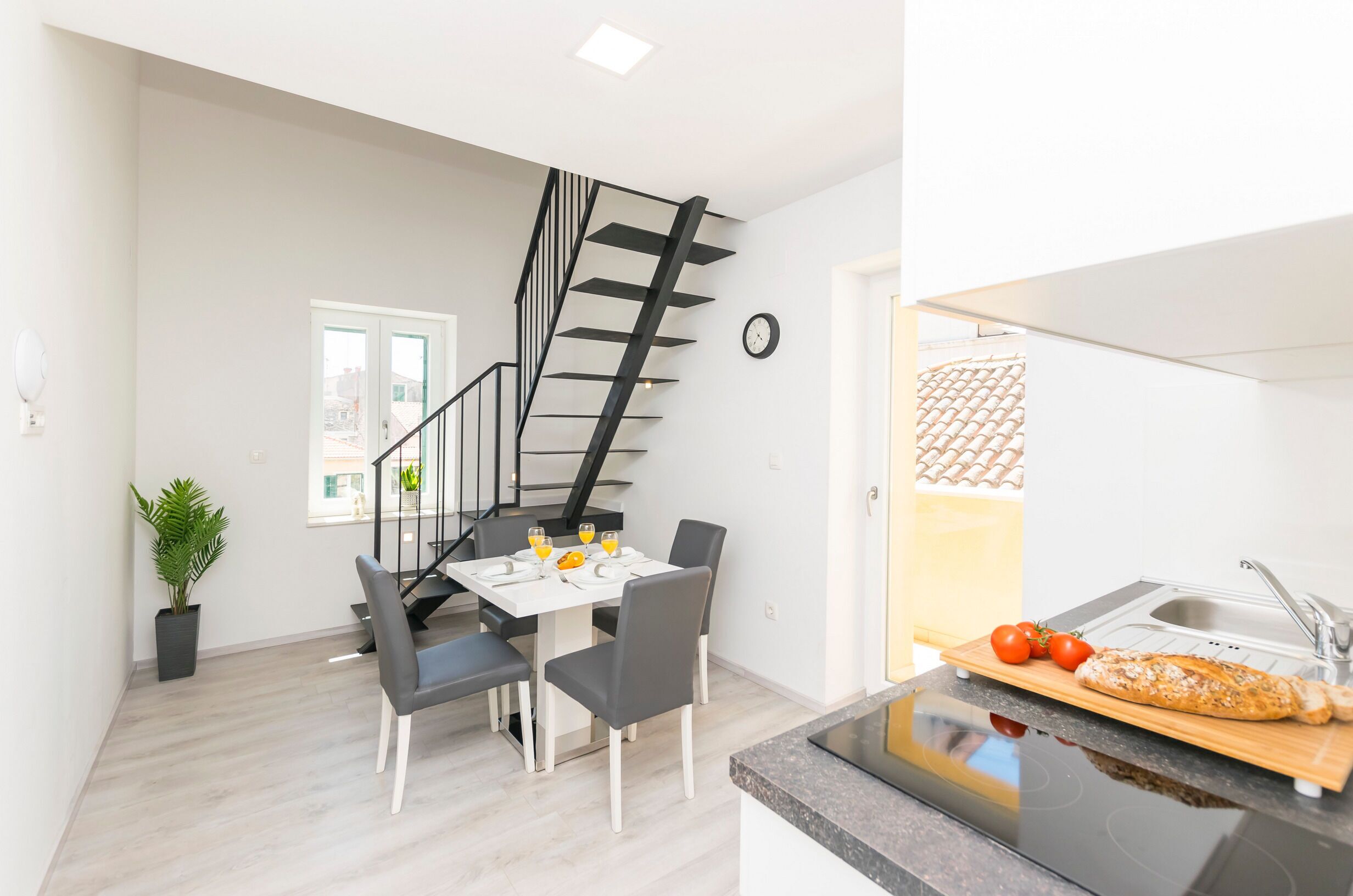A modern kitchen with a built-in oven, a dining area with a table and chairs, and a staircase with a black railing.