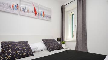 Un dormitorio ordenado con cama, almohadas, una mesita de noche con una planta y una ventana con cortinas.