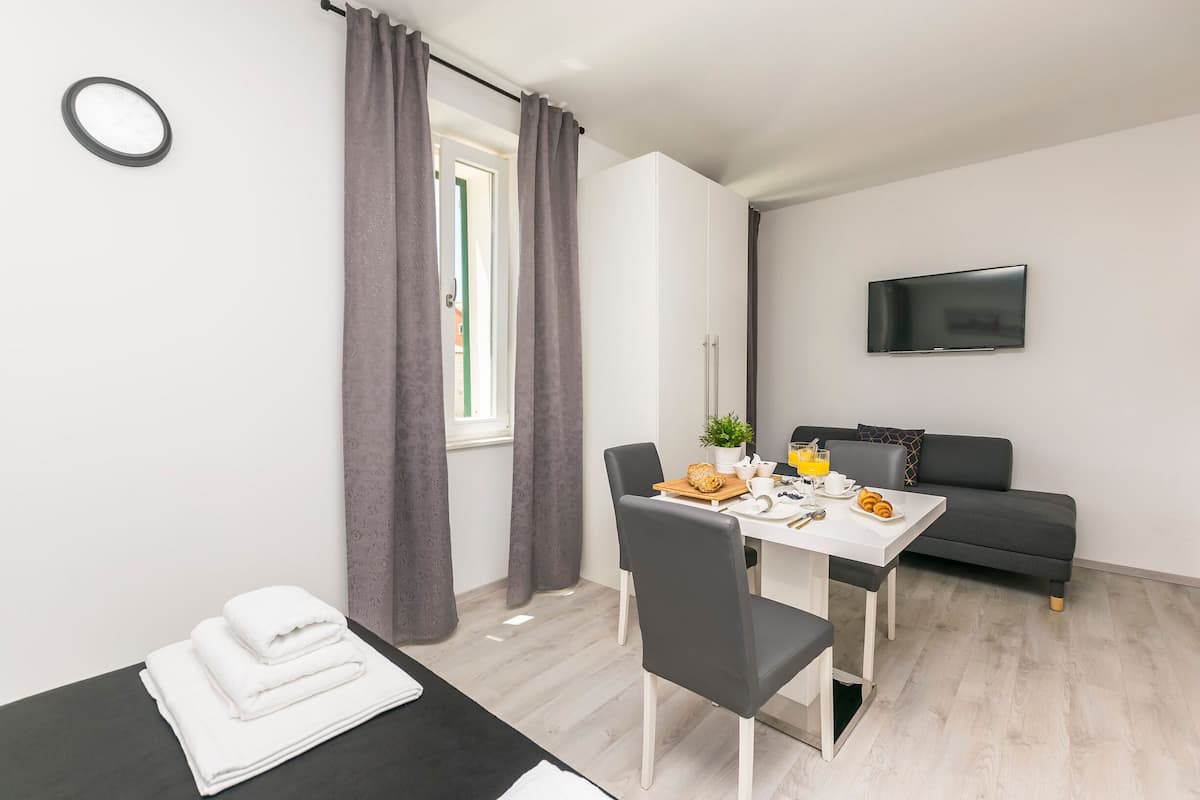 Une pièce moderne et bien éclairée, comprenant une table à manger, des chaises, un canapé, un téléviseur à écran plat et une fenêtre avec des rideaux.