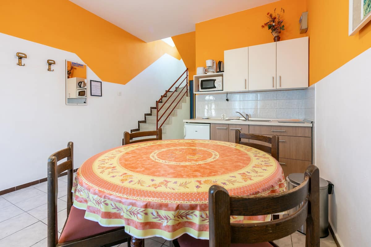 Un coin repas avec une table ronde recouverte d’une nappe à motifs floraux, des chaises en bois et un escalier menant à une cuisine équipée de placards et d’un four à micro-ondes.