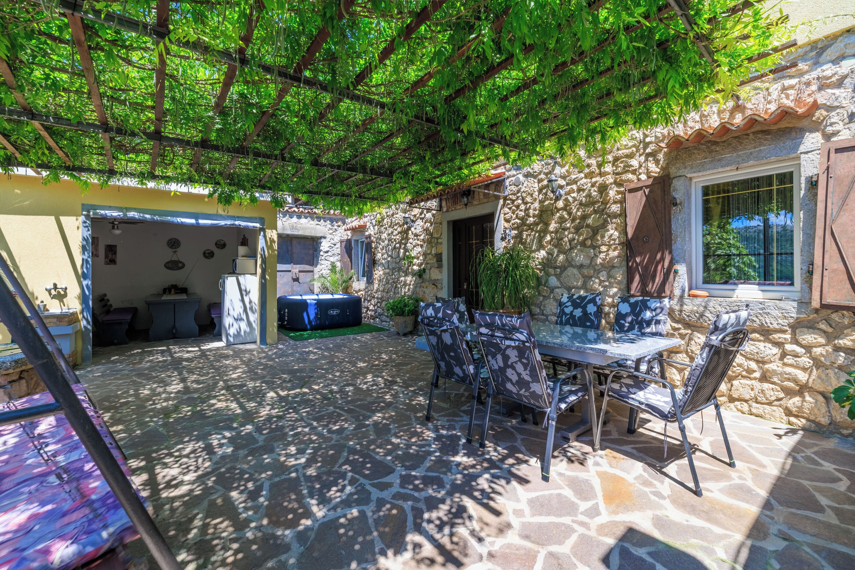 Eine Steinterrasse mit Tisch und Stühlen, eine Pergola mit Grünpflanzen und eine Steinsäule.