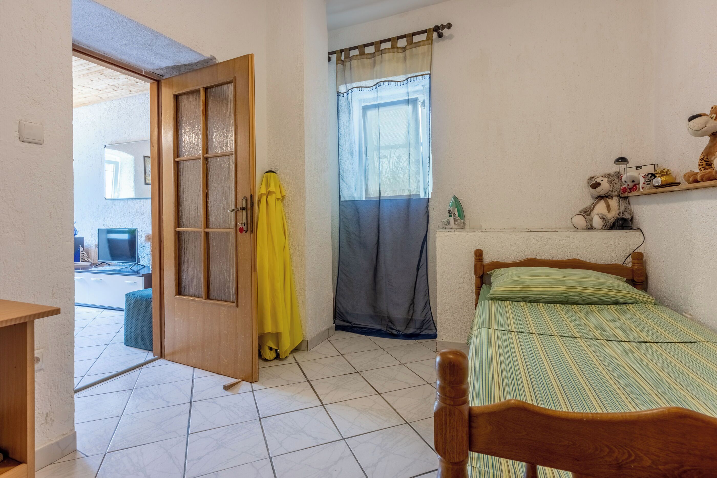 A bedroom with a bed, a wooden door, a window with curtains, and a shelf with stuffed animals.