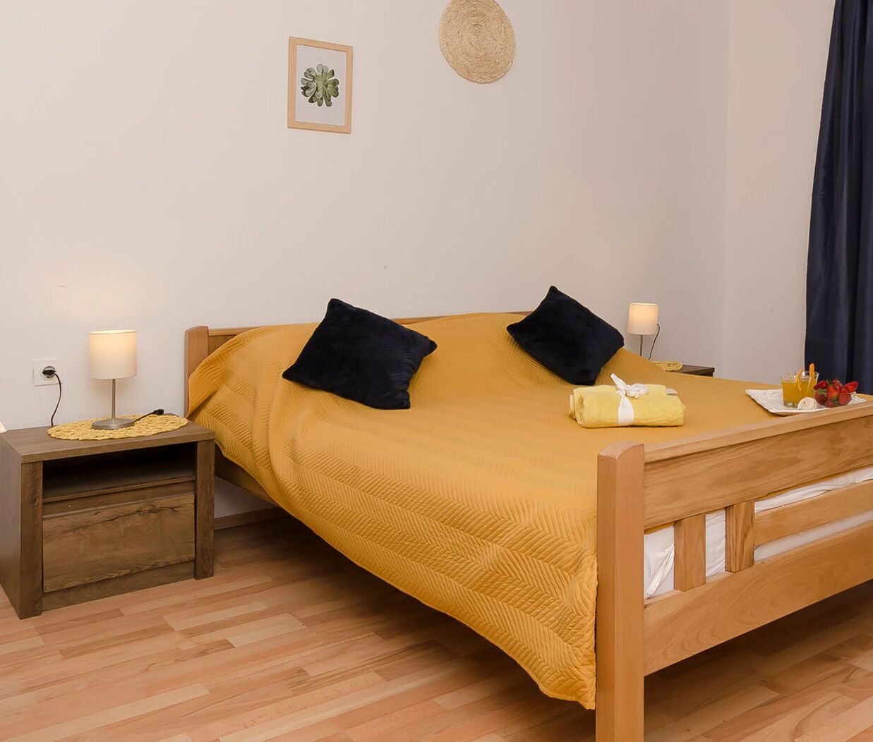 A single bed with a wooden frame, a mustard-colored bedspread, two black pillows, a small bedside table with a lamp, and a wall-mounted decorative item.
