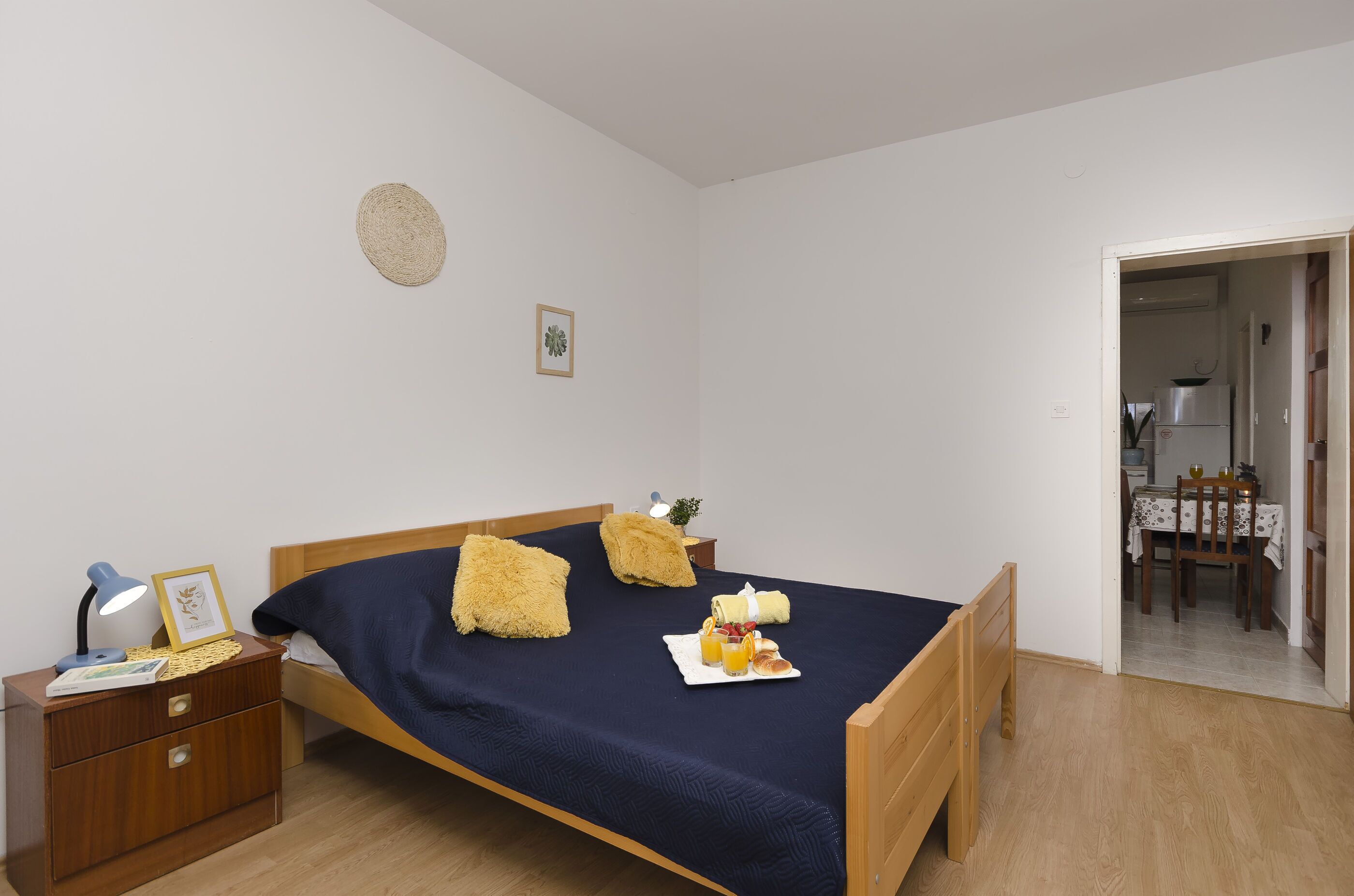Ein ordentlich gemachtes Bett mit einer dunkelblauen Tagesdecke, zwei flauschigen Kissen und einem Frühstücksbrett mit Toast und Saft. Ein Nachttisch aus Holz mit einer Lampe und einem gerahmten Bild steht neben dem Bett.