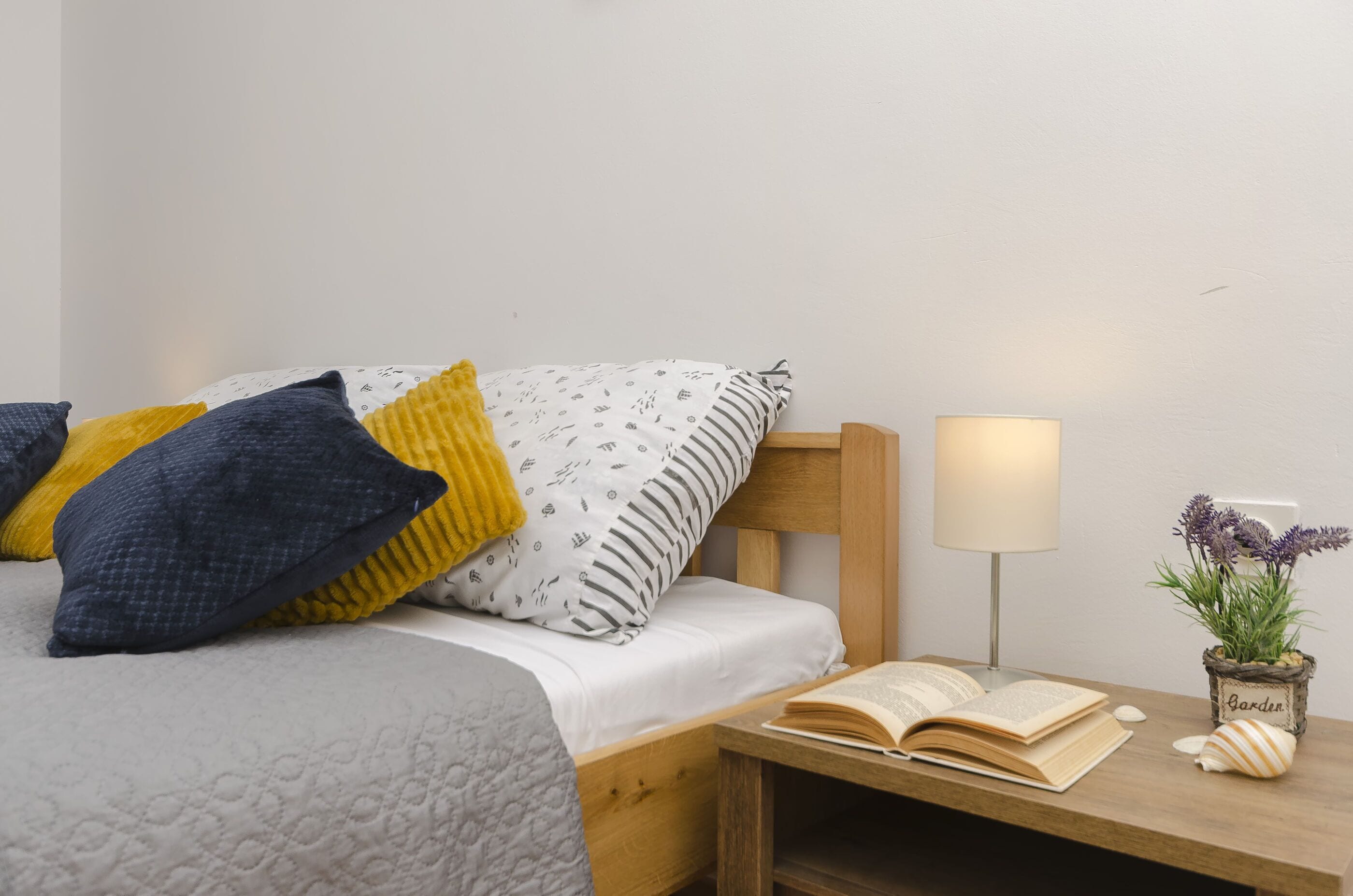 Un lit bien fait, agrémenté d’oreillers à motifs, une table de chevet avec une lampe, un livre ouvert et une petite plante en pot.