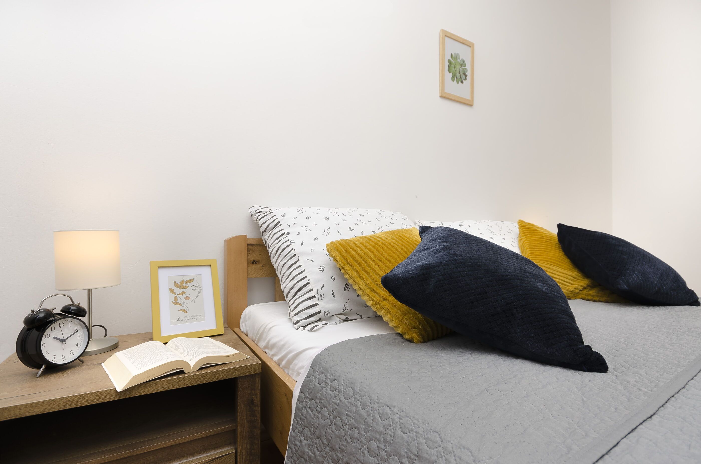 Una cama bien hecha con un edredón gris, dos almohadas (una amarilla y otra azul oscuro), y una mesita de noche con una lámpara, un libro abierto y un cuadro enmarcado.