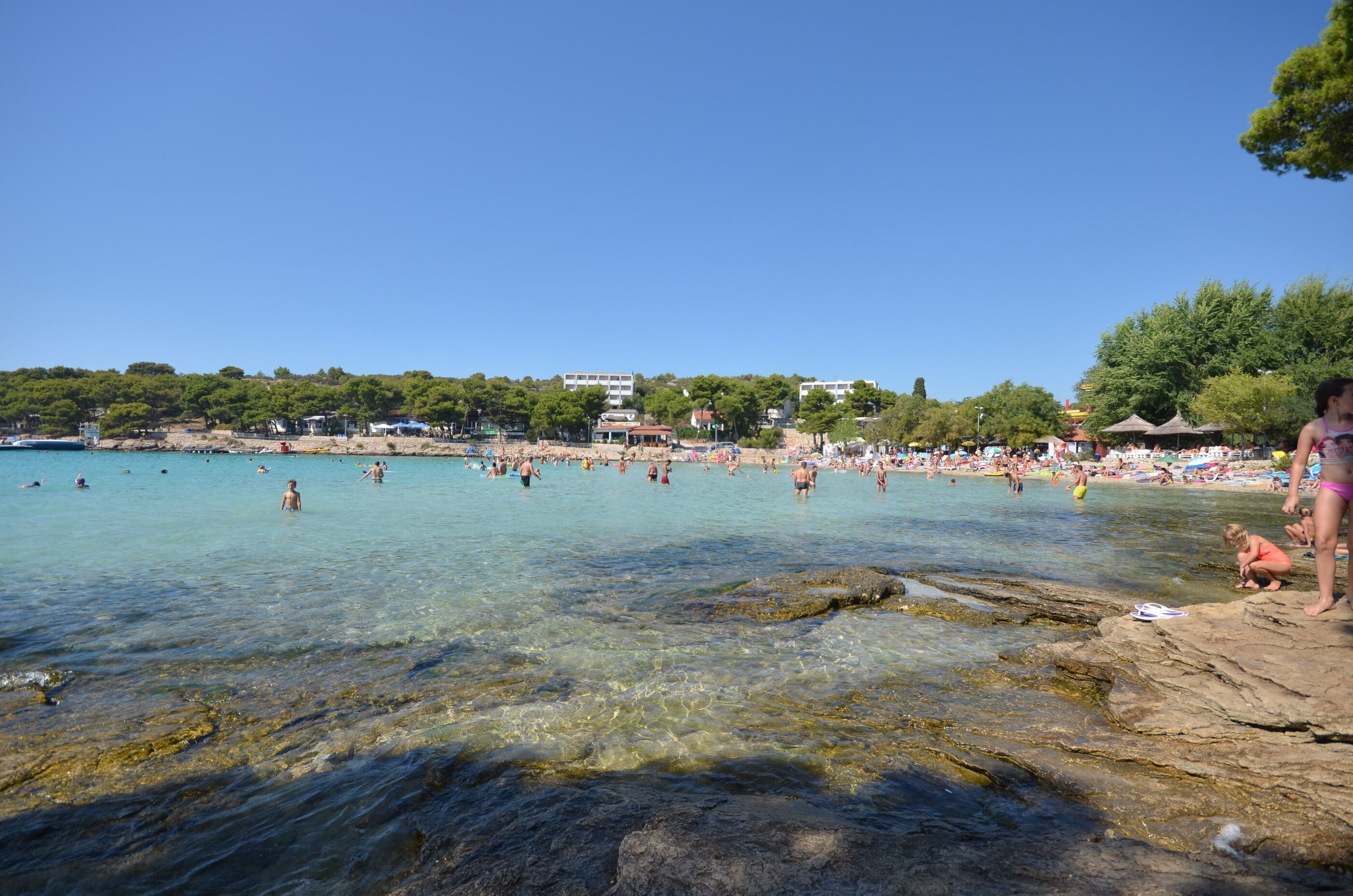 En lys strand med badegæster og solbadere, en klippefyldt kystlinje og bygninger i baggrunden.