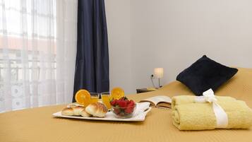 A neatly made bed with a yellow towel and a folded towel with a white ribbon. A tray with croissants, strawberries, and orange slices is on the bed. There is a book and a lamp on a bedside table.