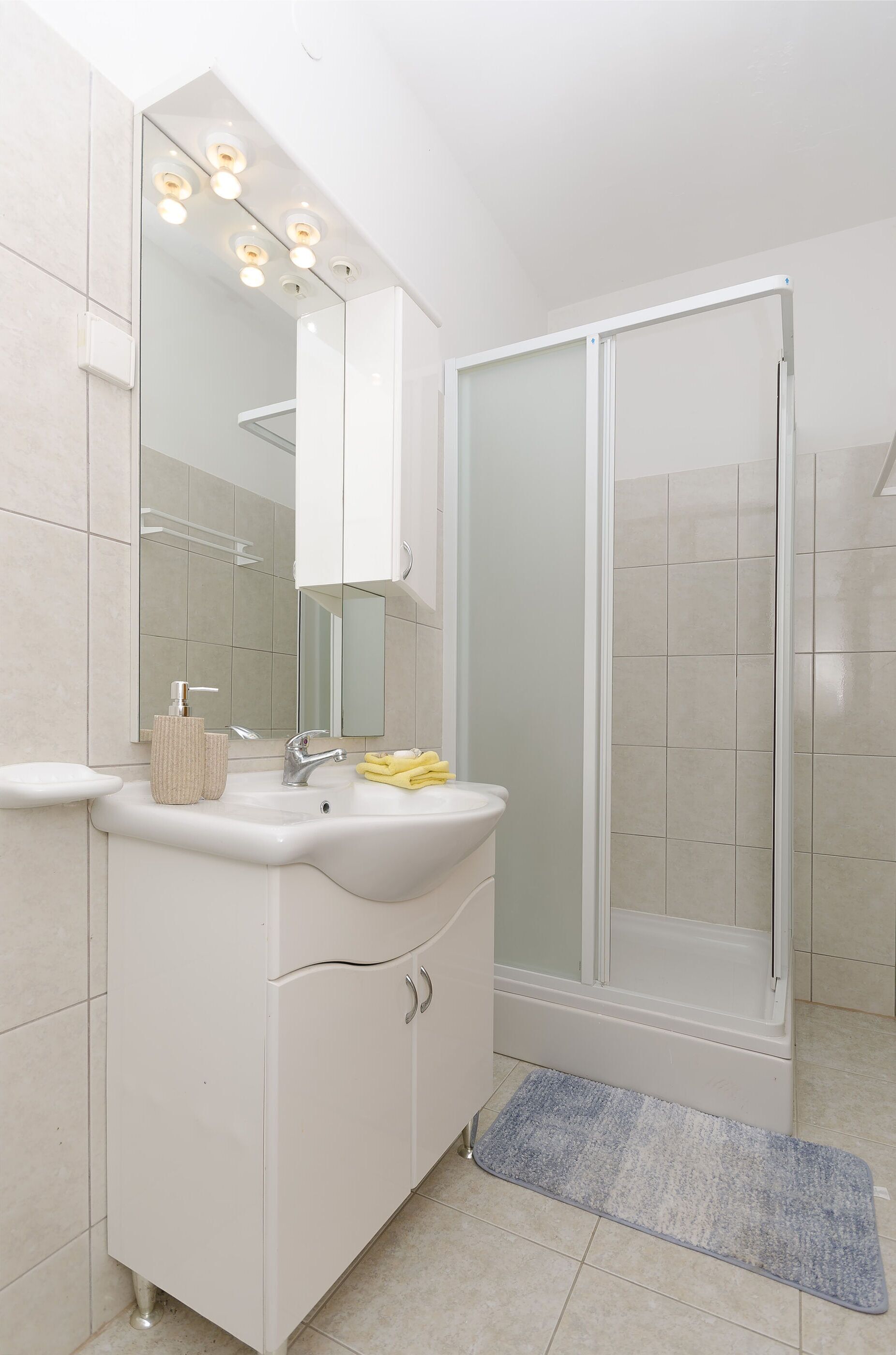 Een moderne badkamer met een witte wastafel, een spiegel met verlichting, een douchecabine en een blauwe badmat.