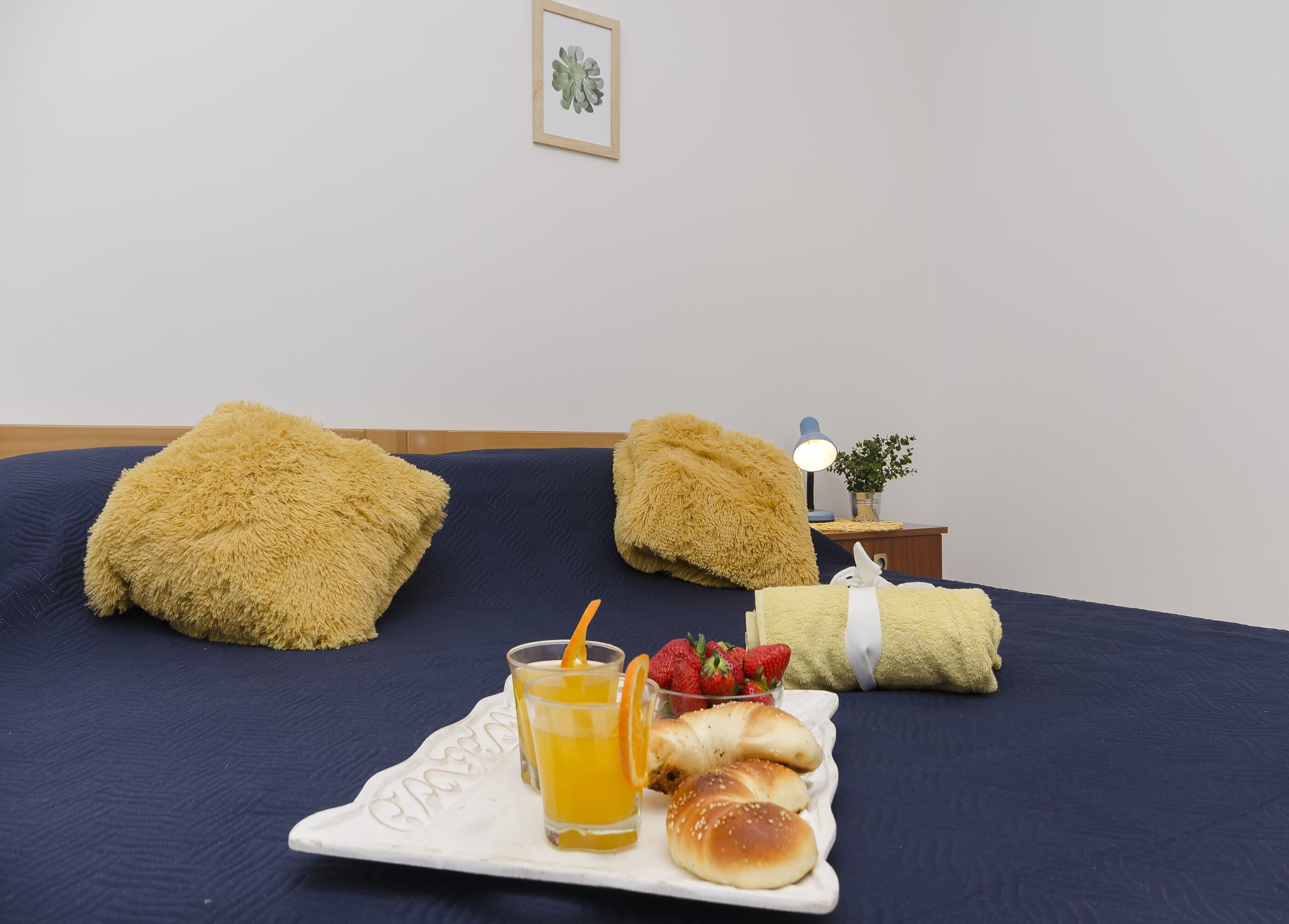 Una cama bien hecha con almohadas mullidas, una mesita de noche con una lámpara, un cuadro enmarcado en la pared y una bandeja con zumo de naranja, fresas y bollería.