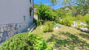 Un bâtiment dont la partie inférieure est en pierre et la partie supérieure est blanche, entouré d’un jardin bien entretenu où l’on trouve divers arbustes et un arbre.