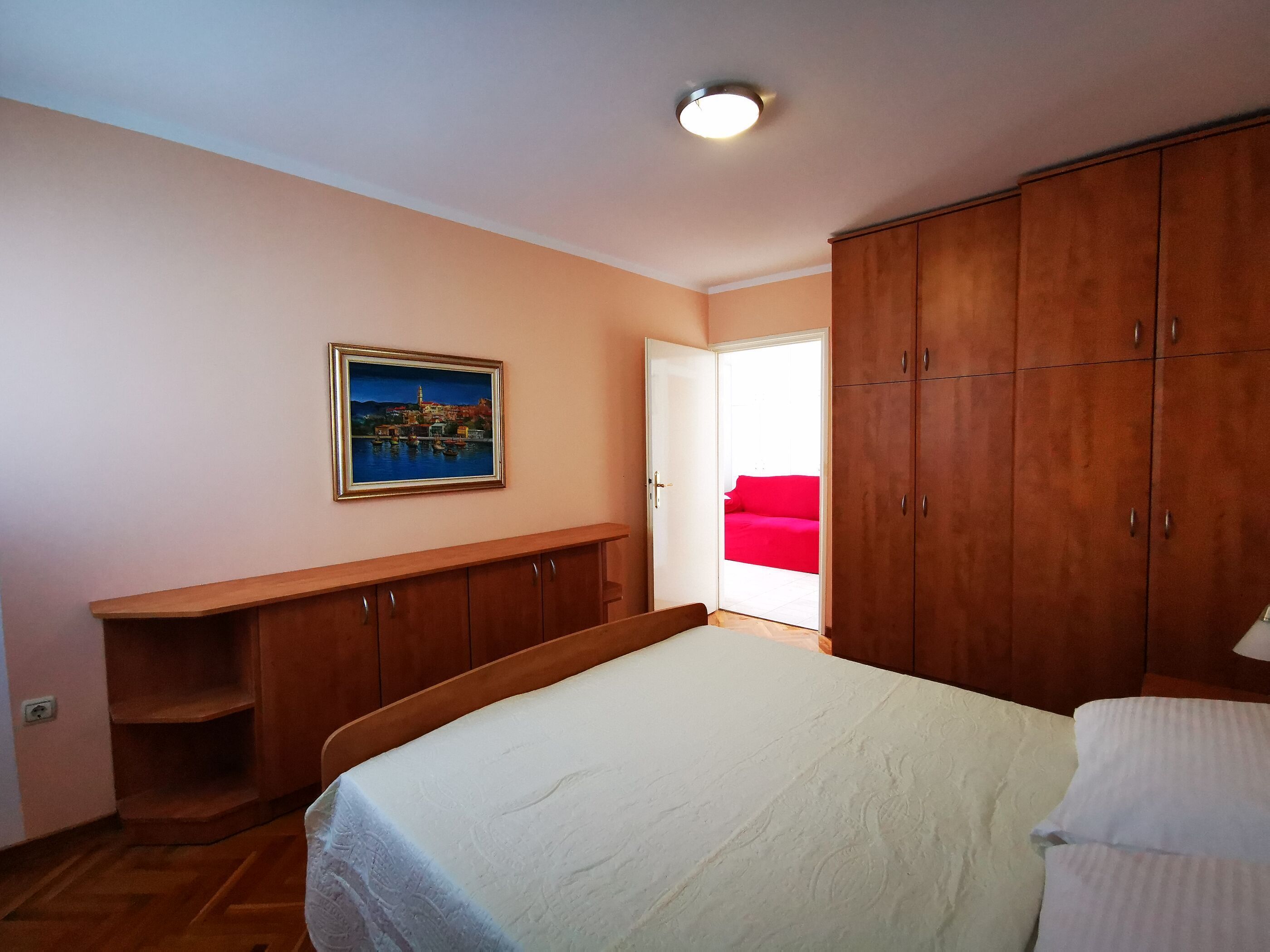 Ein Schlafzimmer mit einem Bett, einem Holzschrank, einem Gemälde und einem roten Sofa im angrenzenden Raum.
