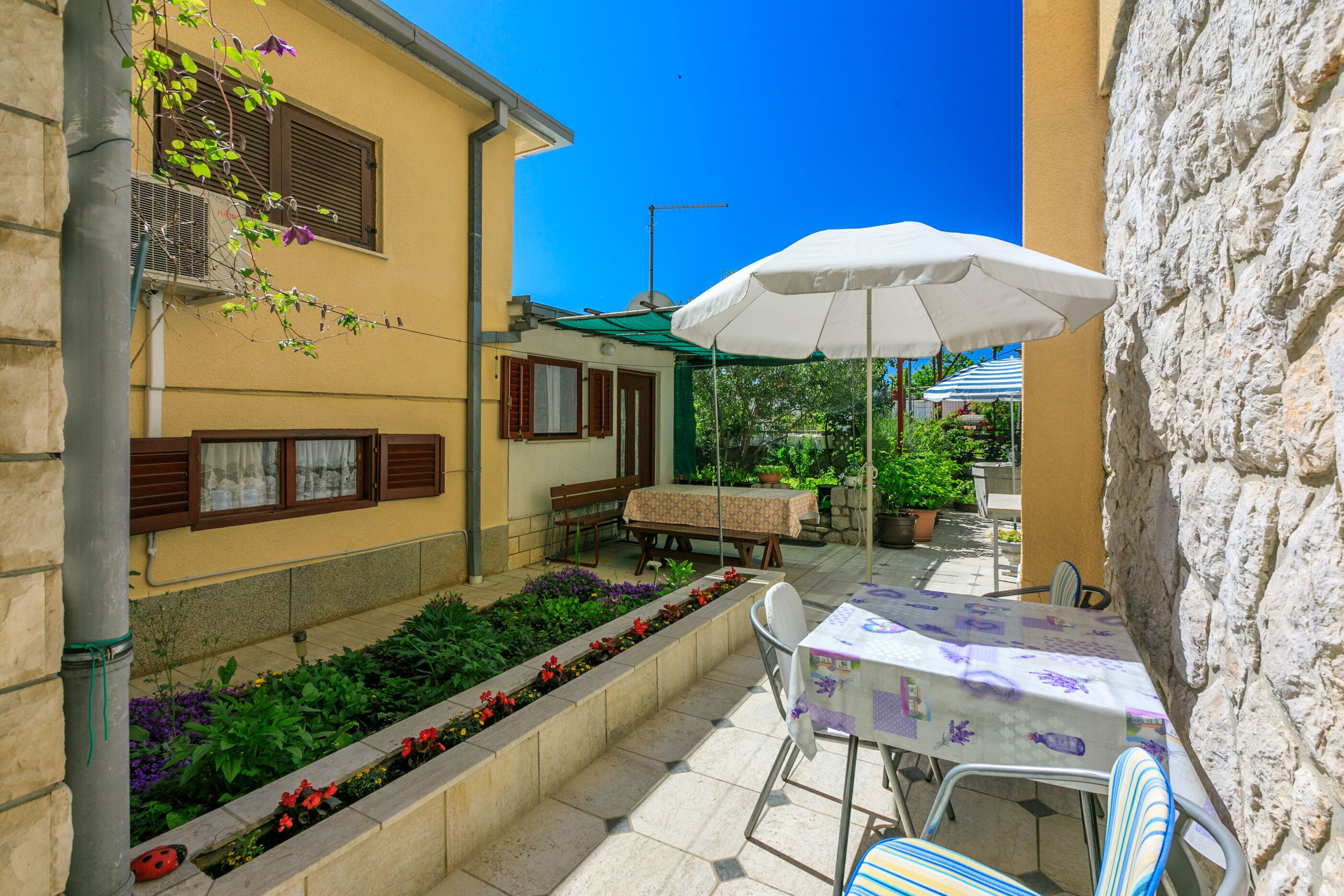 A patio with a table and chairs, a white umbrella, and a flower bed.